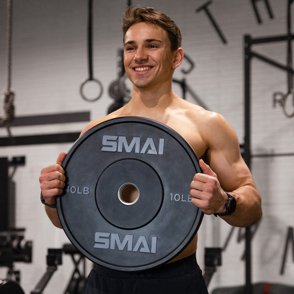 A smiling, shirtless man holds a SMAI USA HD Bumper Plate - (Pair) 10lb in both hands at the gym, with fitness equipment and CrossFit gear in the background.