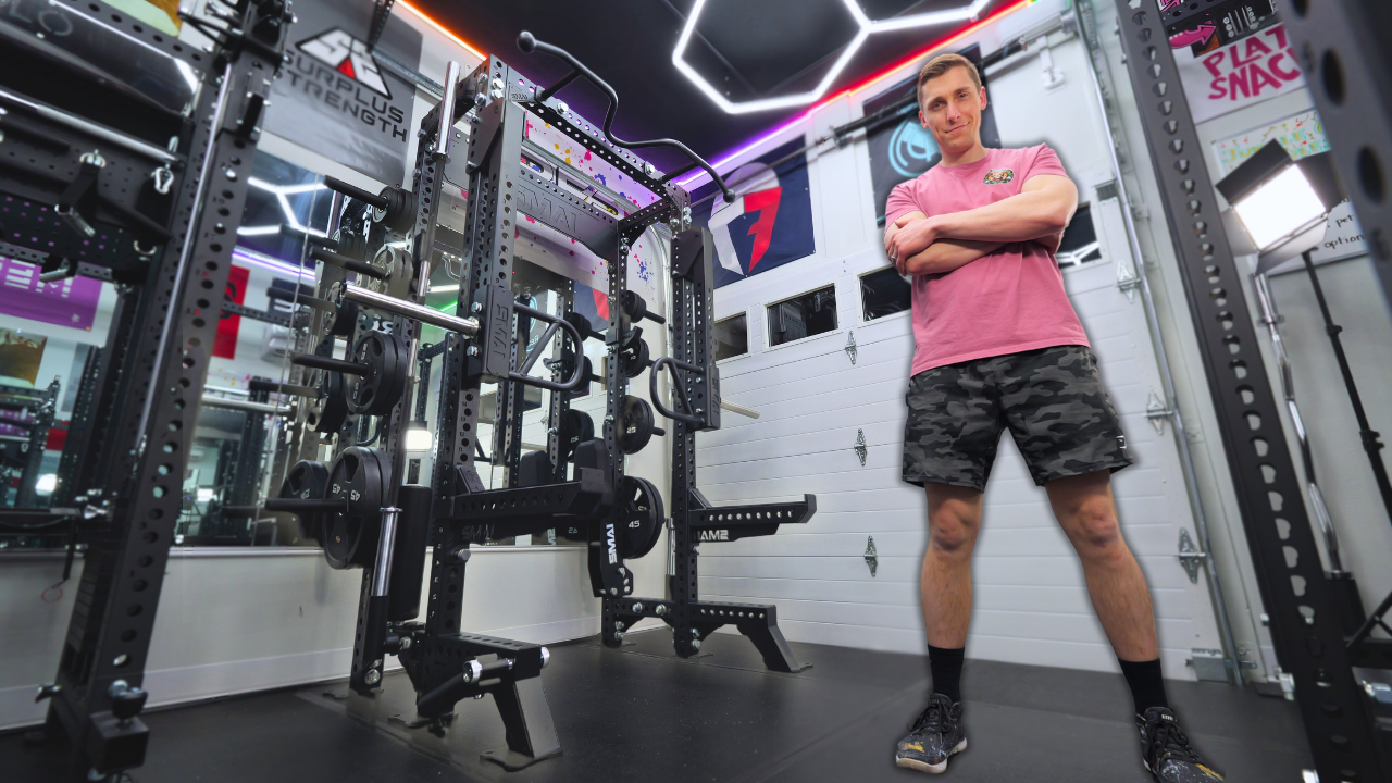 A man in a pink shirt and camo shorts stands confidently with arms crossed in a modern, well-equipped home gym with weights, racks, and colorful LED lighting.