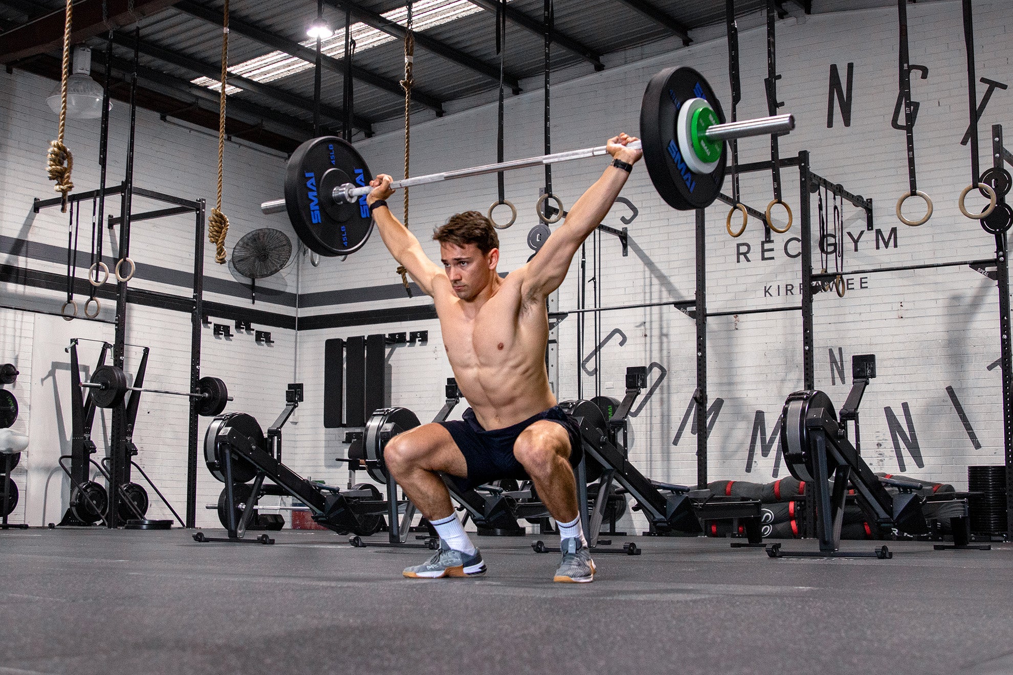 A shirtless man in shorts performs an overhead squat with a loaded barbell in a gym, surrounded by exercise equipment and gymnastic rings hanging from the ceiling.