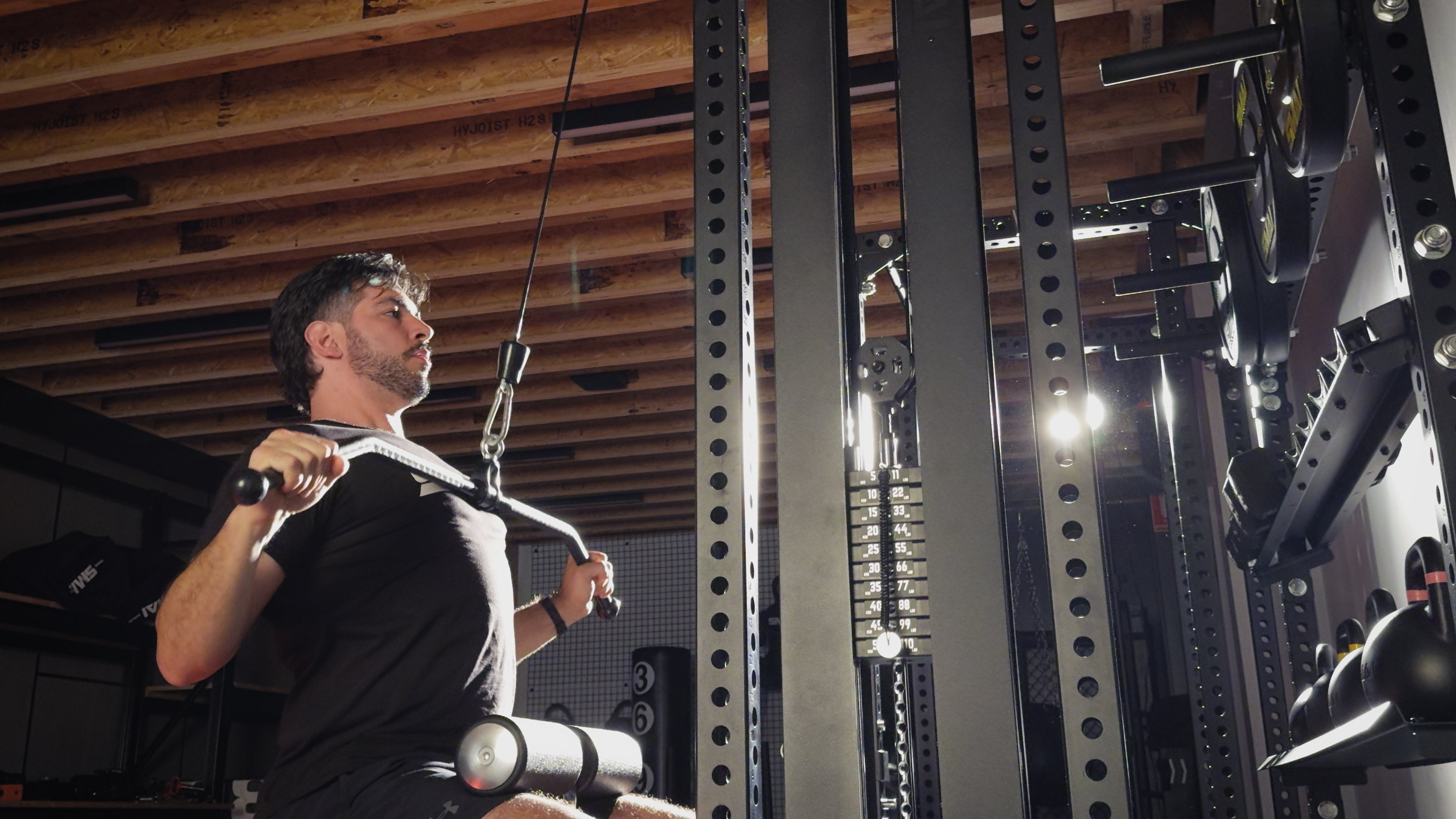 A man in a black shirt performs a lat pulldown exercise on a cable machine in a gym with exposed wooden beams on the ceiling. Bright light shines through the equipment.