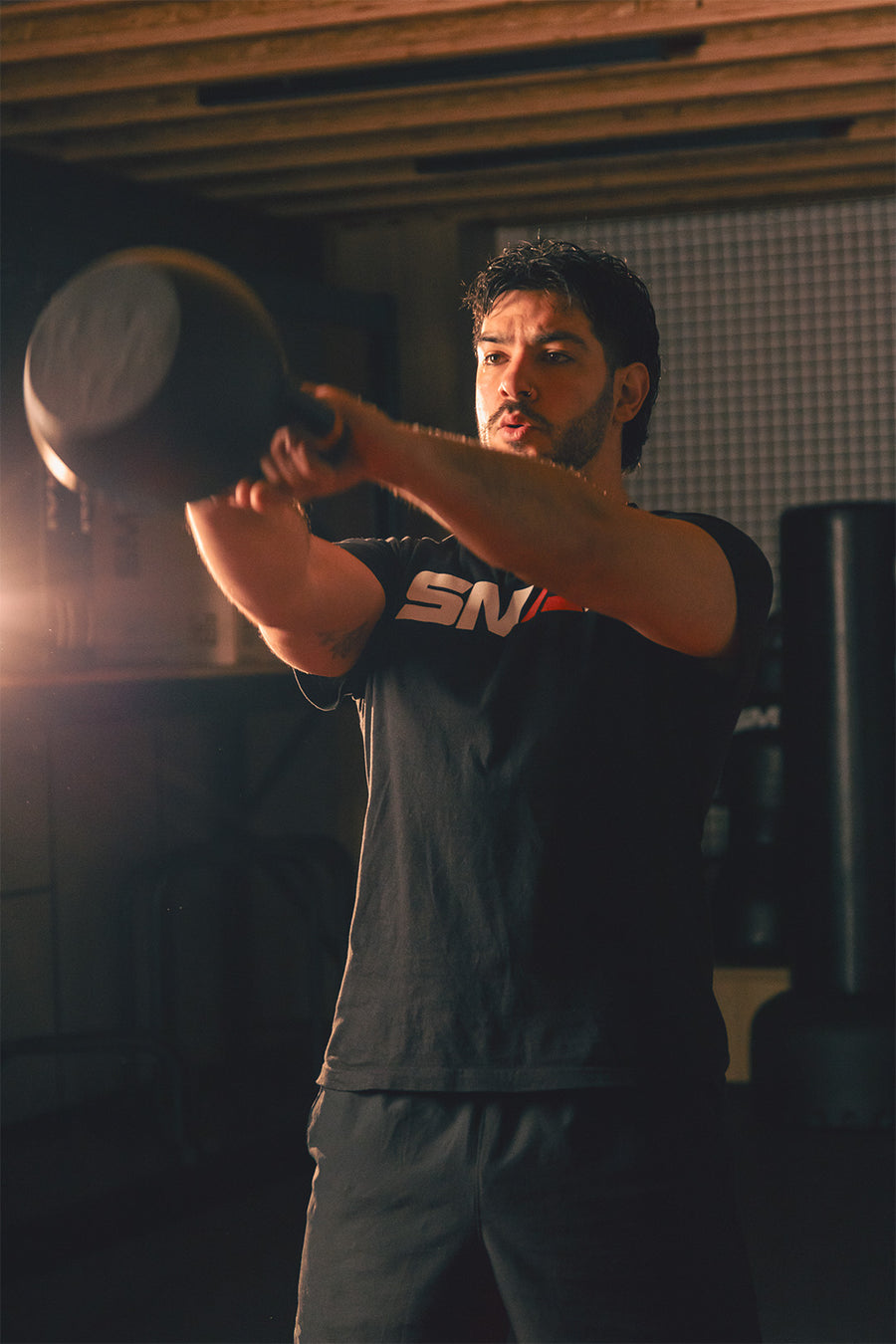 A man wearing a black t-shirt and shorts is lifting a kettlebell with both hands in a gym, with focused expression and warm lighting highlighting his face and arms.