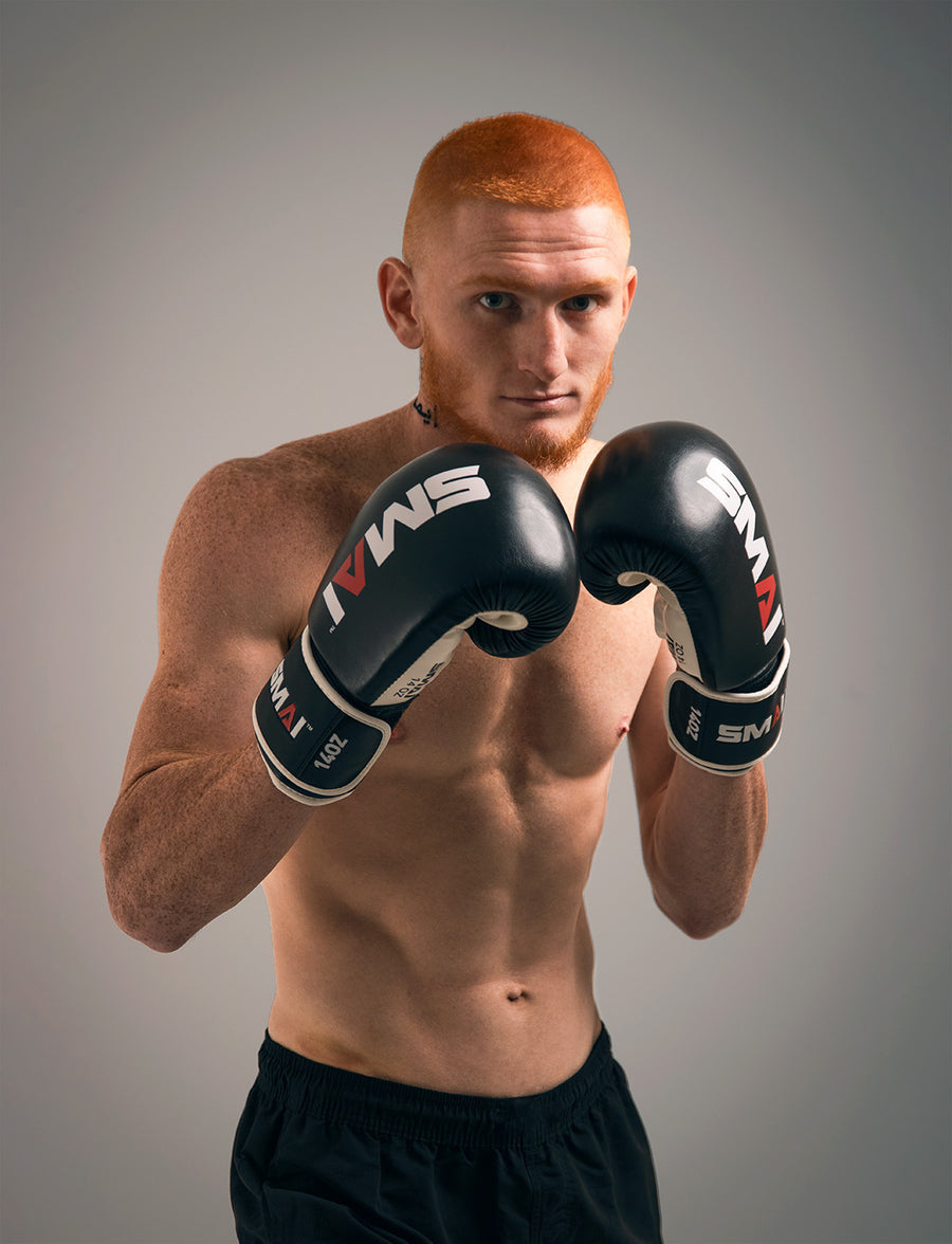 A shirtless man with short red hair wears black boxing gloves and black shorts, standing in a boxing stance with his fists raised, looking confidently at the camera against a plain background.