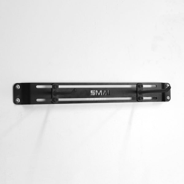 The SMAI USA Workout Mat Wall Storage Rack, made of black metal with a laser-cut "SMAI" logo and mounted by four screws, provides stylish mat storage against a white wall.