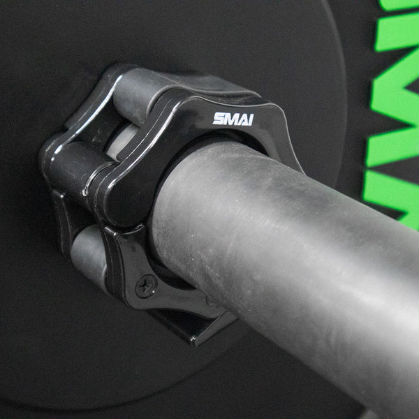 Close-up of SMAI USA Easy Lock Barbell Collars 2" (Pair) securing a weight plate, with the "SMAI" brand visible on the clamp and part of a green logo on the plate in the background. Weight lifting essential.