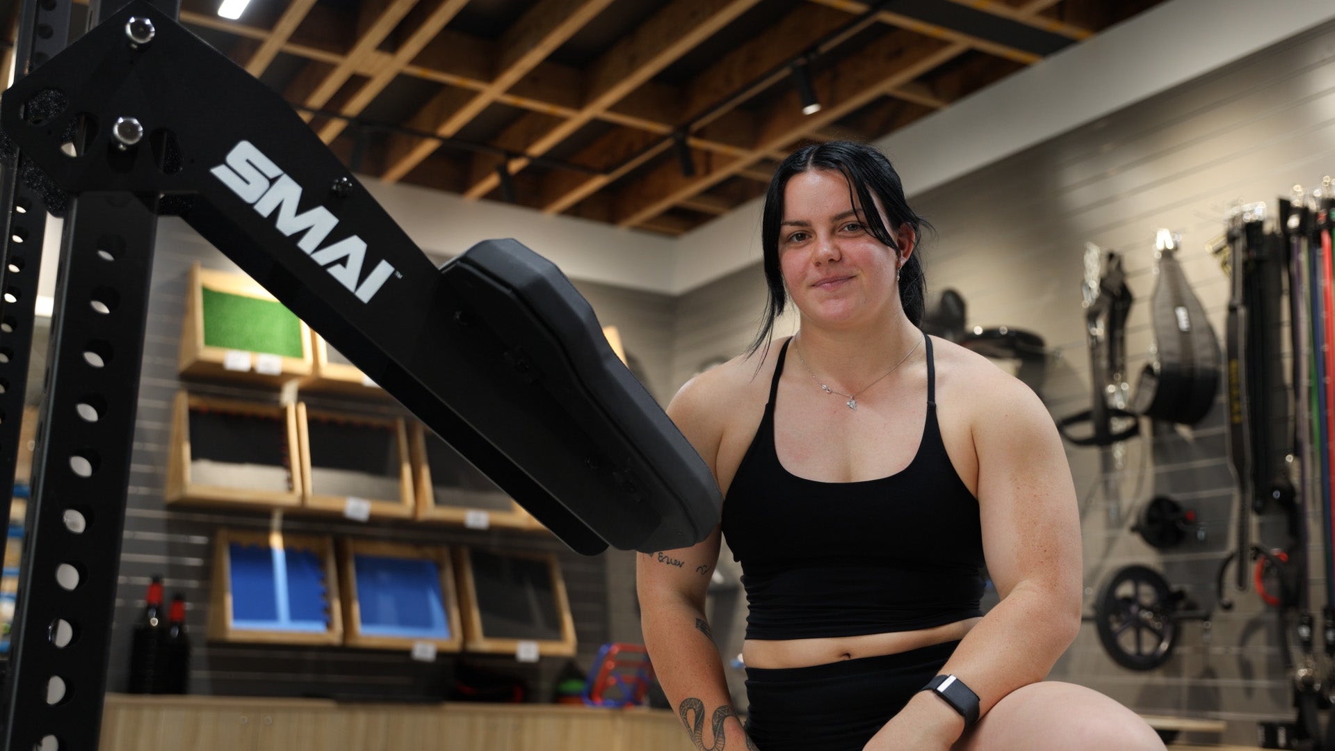 A woman in a black sports bra and shorts sits in a gym, smiling at the camera. Gym equipment and colorful chalkboards are visible in the background. The word "SMAI" is printed on a nearby piece of equipment.