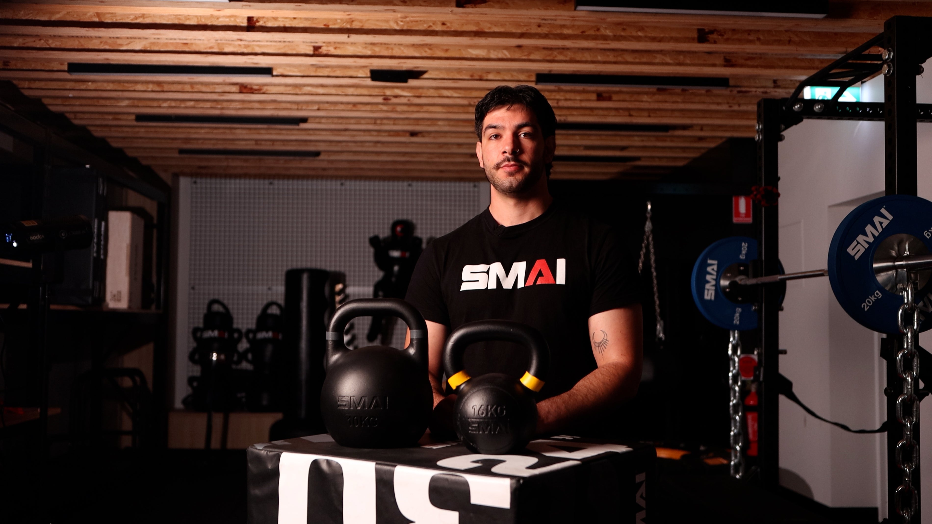 A man with a mustache, wearing a black "SMAI" t-shirt, stands in a gym behind two kettlebells placed on a black and white box. Gym equipment is visible in the dimly lit background.