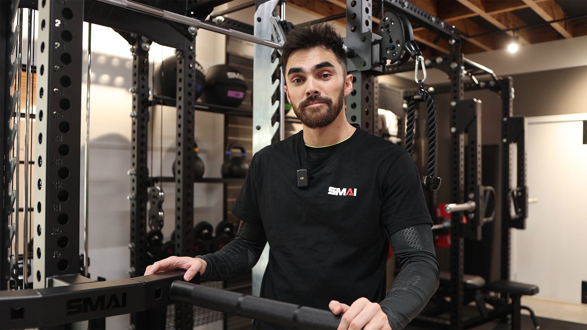 A man with short dark hair and a trimmed beard stands in a gym, wearing a black T-shirt and sports gear. He holds onto gym equipment, with weight racks and fitness gear visible in the background.