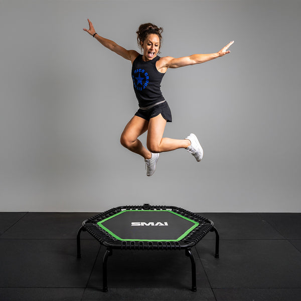 A woman in black activewear and white sneakers jumps joyfully on the SMAI USA Rebounder Mini Trampoline 50" in a studio with a gray background.