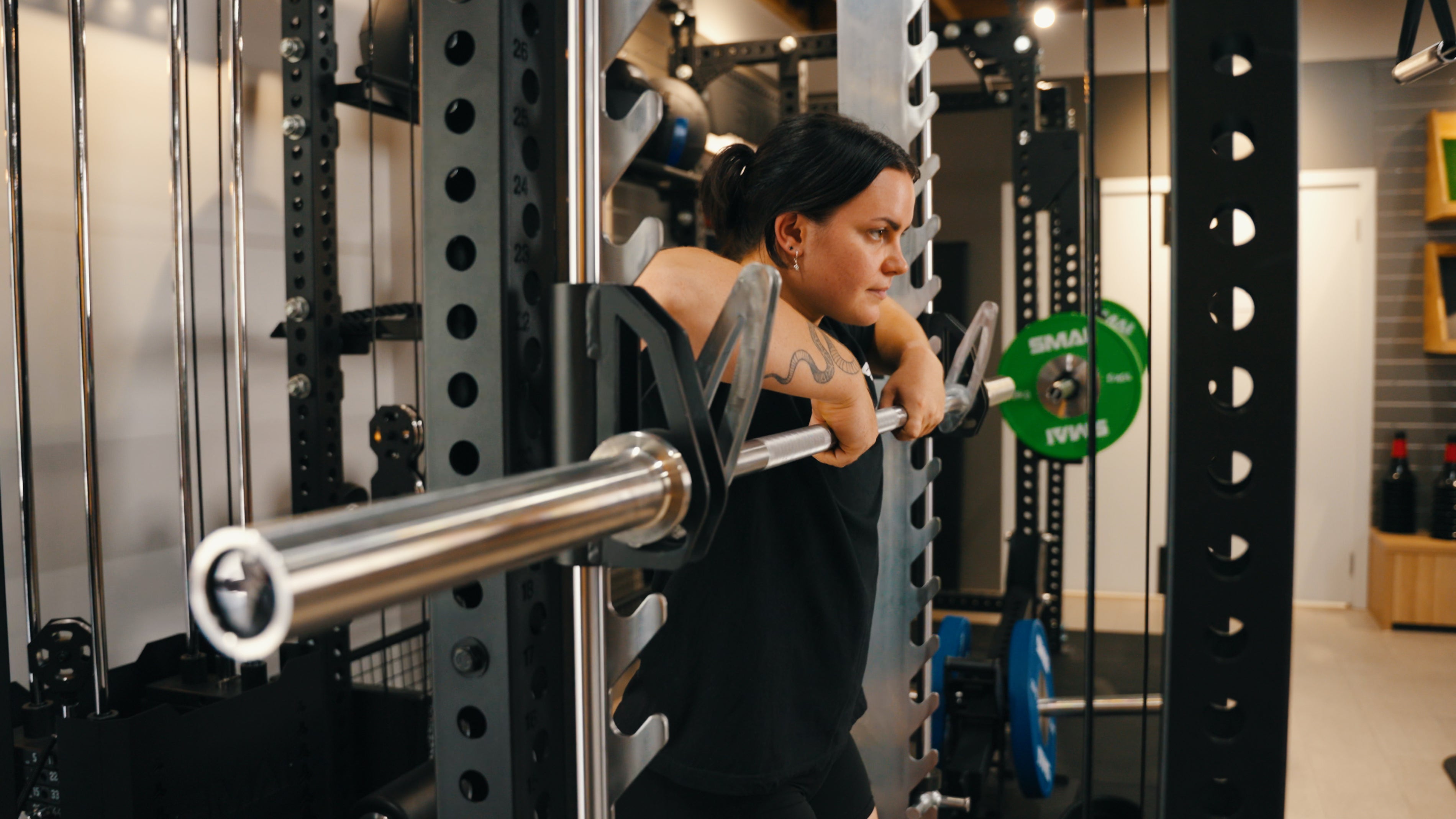 A person with dark hair and a visible arm tattoo performs an upright row with a barbell in a gym, using a power rack and surrounded by weightlifting equipment.