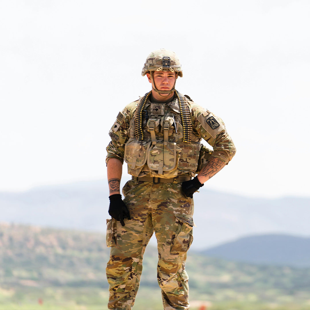 A soldier in full camouflage uniform and helmet stands outdoors with a serious expression. He has ammunition draped over his shoulders and wears black gloves amid a mountainous landscape in the background.