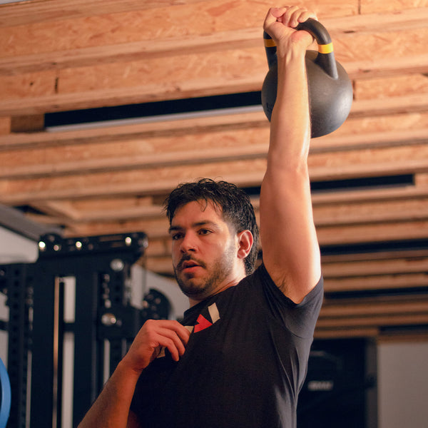 A man in a black shirt lifts a SMAI USA Competition Kettlebell with a large handle overhead in a gym with exposed wooden beams, looking focused on his workout.