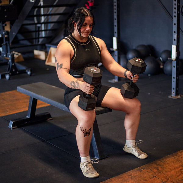 Wearing athletic gear, a person sits on a gym bench holding SMAI USA Rubber Hex Dumbbells (Pair), ready to work out. The gym includes black walls, various equipment, and stairs visible in the background.