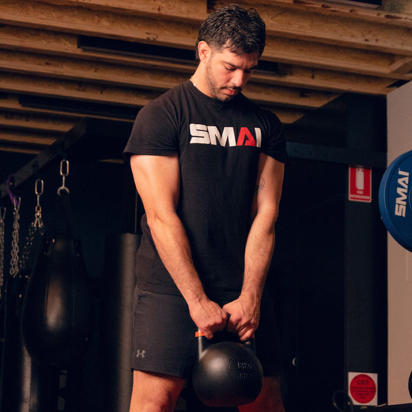 A man in a black SMAI USA shirt and shorts stands in a gym under a wooden ceiling, holding SMAI USA Competition Kettlebells with both hands. Gym equipment is visible in the background as he looks down at the kettlebell.