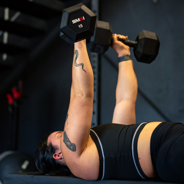 A person in athletic wear lies on their back lifting two SMAI USA Rubber Hex Dumbbells (Pair) above their chest in a gym. Their tattooed arms and commercial grade gym equipment are visible in the background.