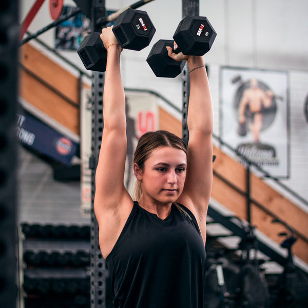 female lifting two 25lb rubber hex dumbbells over head in a gym