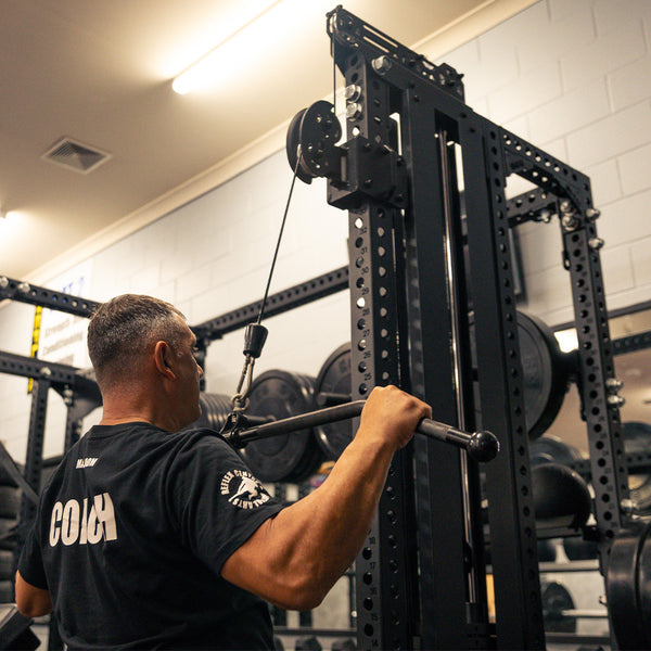 A person in a black shirt uses the SMAI USA Lat Pull / Low Row Cable Tower Add On - Vanta Series for a lat pulldown exercise, with weight plates and other gym equipment visible in the background.